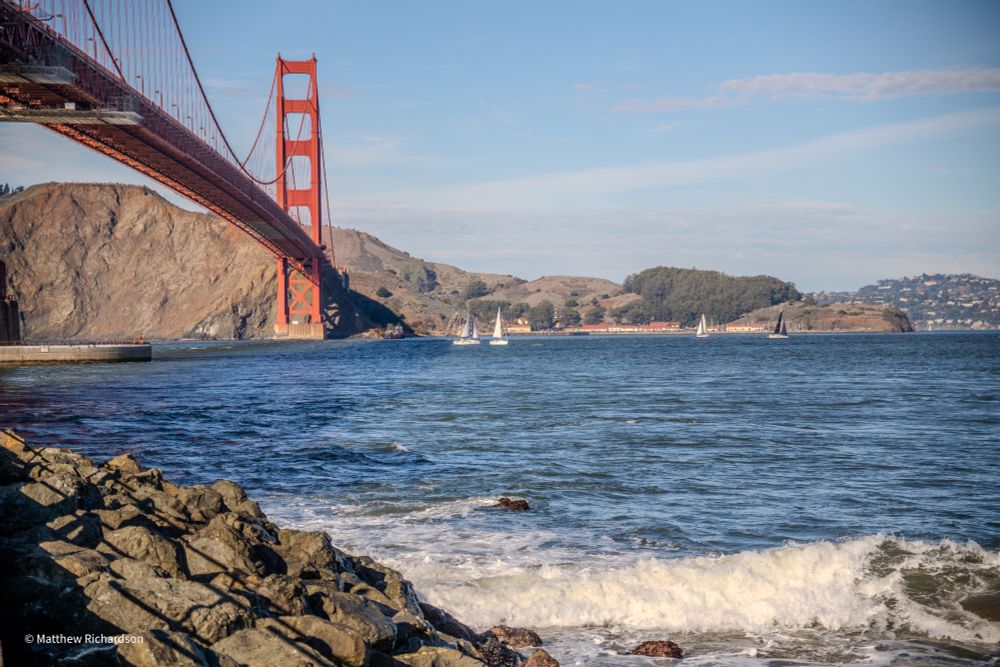 Golden Gate Bridge pictured on the left with sail bots in the far background and crashing waves up close. 