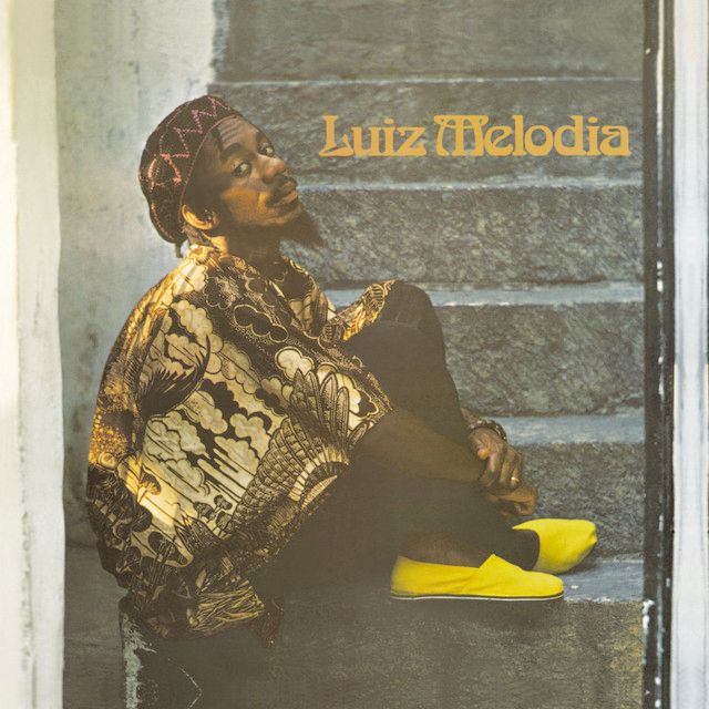 capa de álbum do cantor luiz melodia. ele está sentado em uma escada de degraus cinzas, de lado, mas olha para a câmera. veste chapéu e camisa estampados em dourado e preto, calça preta e sapato estilo alpargata amarelo neon. 