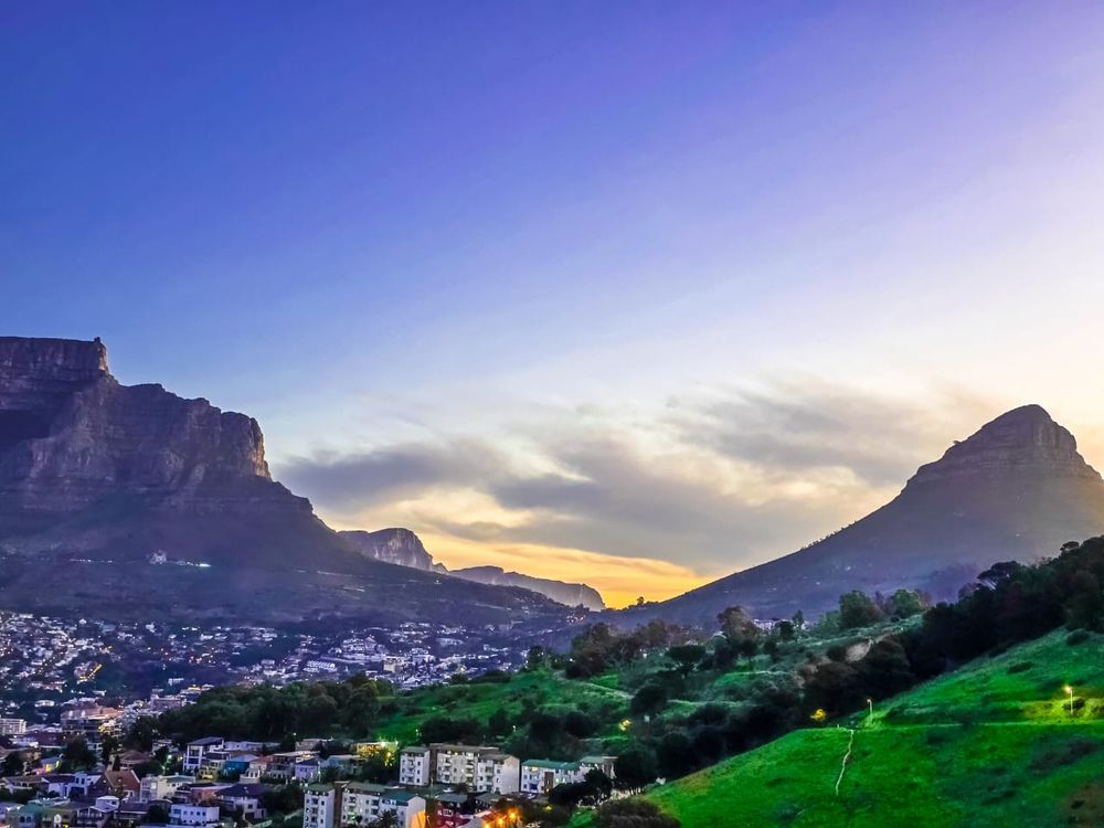 A wide view of Cape Town at sunset, framed by two iconic mountains. On the left rises the flat-topped Table Mountain, while on the right stands the conical peak of Lion’s Head. Below them, the city stretches out with clusters of houses and buildings nestled among green hills. The sky glows in soft shades of blue, purple, and gold as the sun dips behind the mountains, casting a tranquil light across the landscape.
Photo by Lynne Quick.