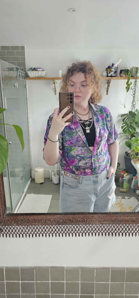 A bathroom mirror selfie of an outfit consisting of high-rise denim shorts and a loose thrift-shop floral silk shirt open to the navel over a sports bra, with a chain and necklace combo