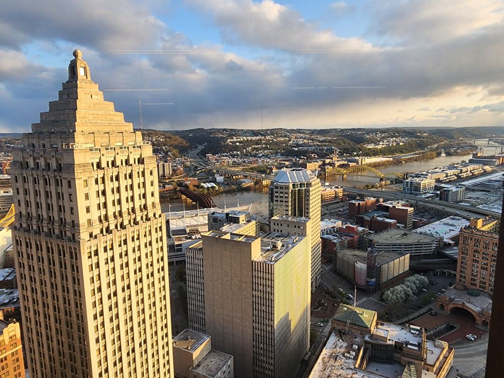 The sky is sunny with some cloud coverage, with hints of blue peaking through. The picture is taken from the north facing side of the U.S. Steel Tower. A small portion of Downtown Pittsburgh is seen, including the Gulf Building. It also overlooks the Strip District and in the distance other neighborhoods across the Allegheny River.
