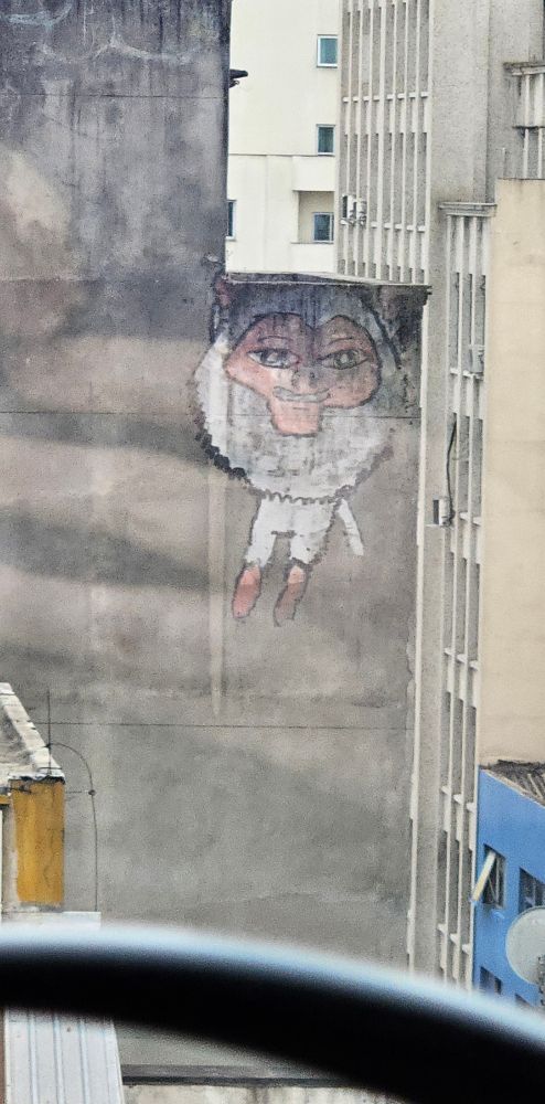 Foto de um pixo numa lateral de um prédio de um desenho de um bicho peludo bípede, com o rosto e os pés cor de rosa e um rabo branco