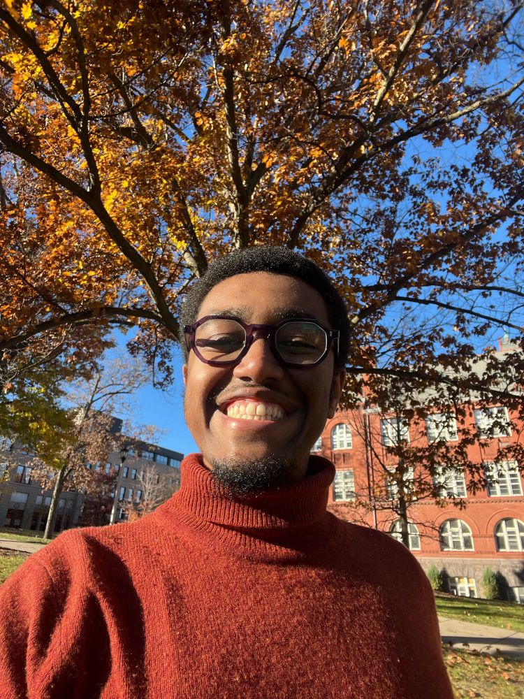 A Black man wearing glasses and a burnt orange turtleneck is smiling in front of a large tree that has turned orange and gold from the autumn weather. The man has short black hair and a small black goatee. The angle of the photo makes it look like the branches of the tree are coming out of his head.