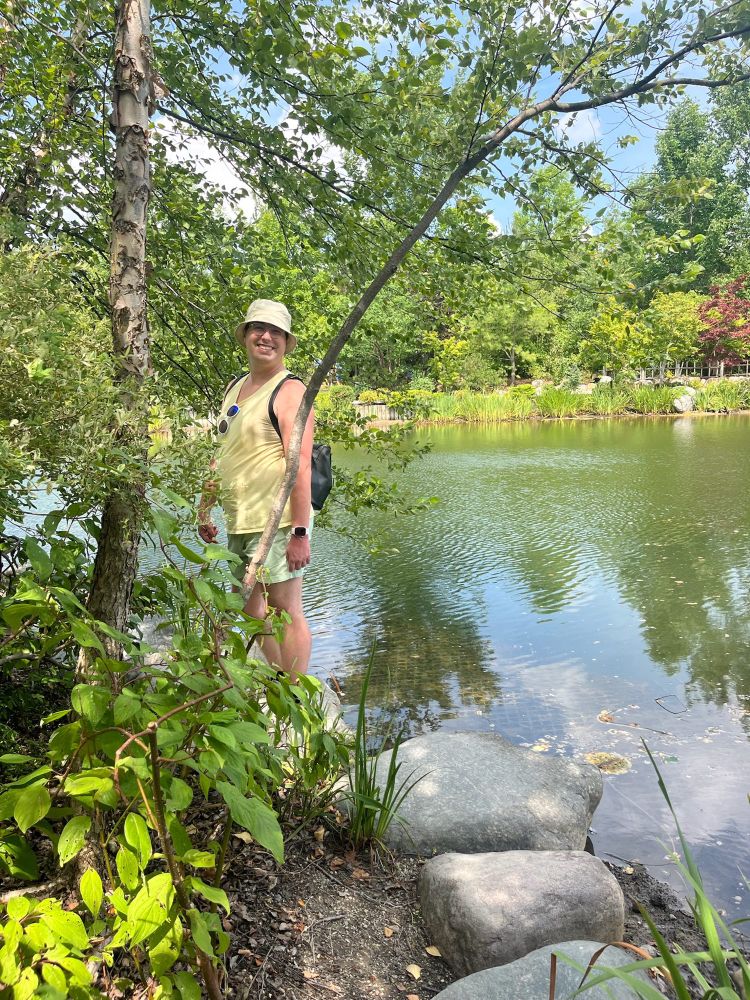 Queer person in a yellow tank top and green shorts by a lake in a garden 