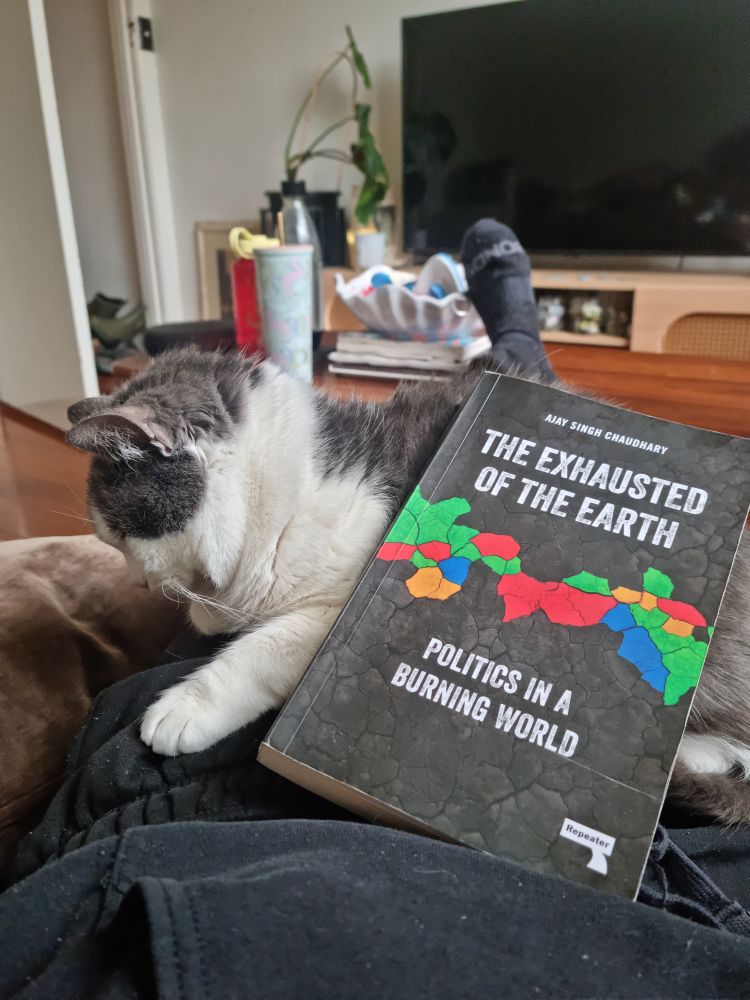 a grey and white cat called Charlie sits on a person's lap. resting on Charlie is a copy of 'the exhausted of the earth' by Ajay Singh Chaudhary.