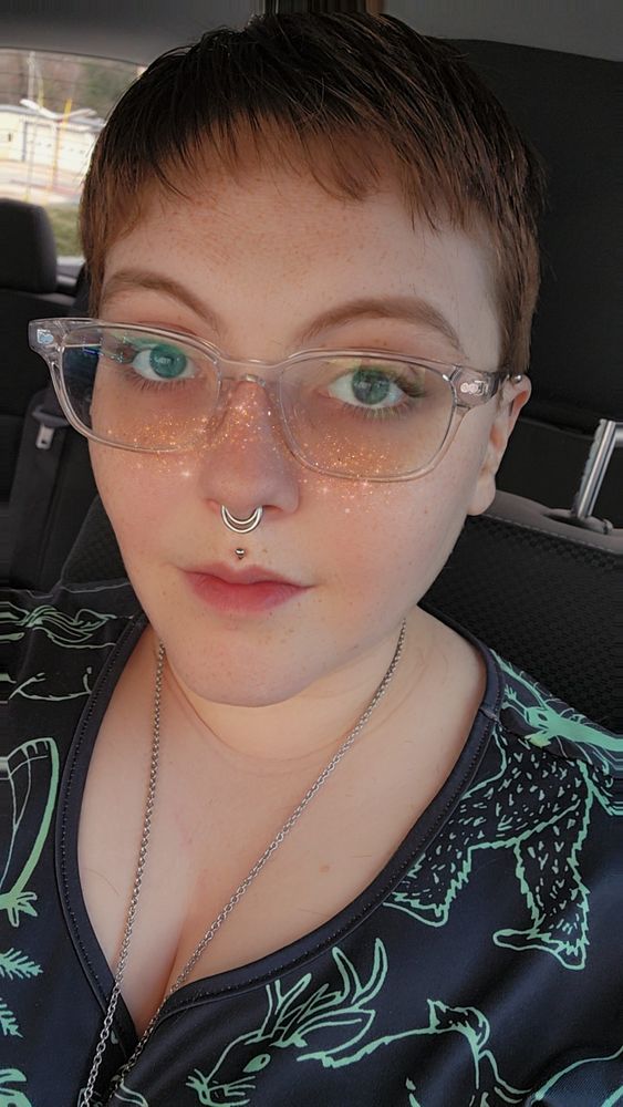 A close-up portrait of a person with short red hair, wearing clear glasses, a septum piercing and a Medusa piercing. The individual has pale skin with freckles and blue eyes and is dressed in a black dress featuring green  cryptid illustrations. The background indicates they are in a vehicle, with soft lighting highlighting their features. There is a snapchat filter on the image giving them sparkle freckles.
