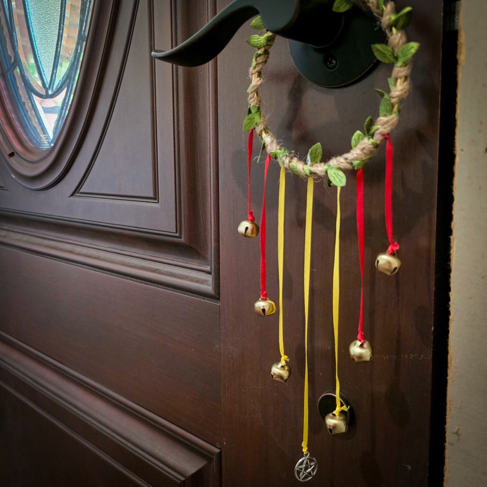 A set of witch bells hangs on a doorknob. The hoop is wrapped with jute twine with green artificial leaves on it. 2 red ribbons, then 3 yellow ribbons, then 2 more red ribbons, all with bells on the ends, hang in a stacked fashion with the center ribbon being the longest and sporting a pentagram charm instead of a bell, representing a flame for Beltane.