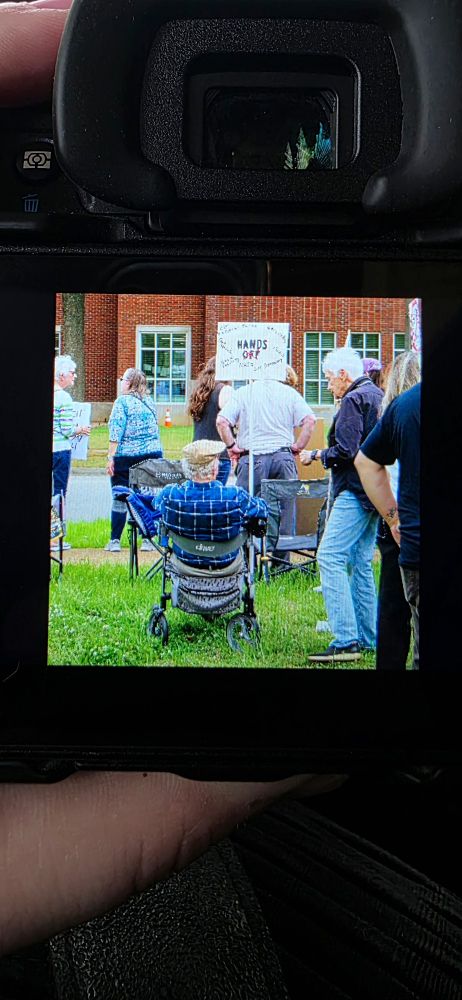 A photo of the back of my camera. The screen shows a portrait photo from behind of a senior citizen in a wheelchair, wearing a blue plaid shirt, holding an illegible protest sign.