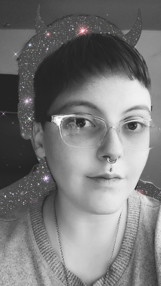 A close-up black and white portrait of a person with very short hair, clea glasses, a septum piercing, and a Medusa piercing. They are wearing a grey sweater. The snapchat filter on the image creates a silhouette behind the person filled with stars and glitter and has playful devil horns. 