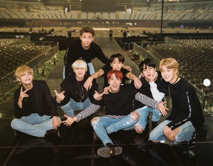 Photo of BTS on stage in an empty arena during the wings tour. They're in light blue jeans and black shirts. 