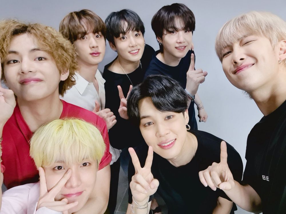 BTS is posing for a selfie with happy expressions in front of a plain gray wall. V, Suga, and RM are holding up their index finger in a "1" pose. J-hope, Jungkook, and Jimin are folding up peace signs. Jin is holding up a thumbs up.