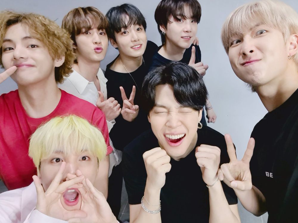 BTS is posing for a selfie with happy expressions in front of a plain gray wall. V and Suga are holding up their index finger in a "1" pose. Jungkook and RM are folding up peace signs. Jin is holding up a thumbs up. J-hope has his hands in front of his mouth in an o-shape shouting out and Jimin has his eyes squeezed shut with a big smile and hands in two fists, cheering.