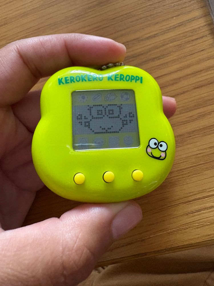 A picture of a hand holding a Kerokeroppi virtual pet toy. The toy is bright green and shaped like a frog head. A girl frog with pigtails is shown on the screen.