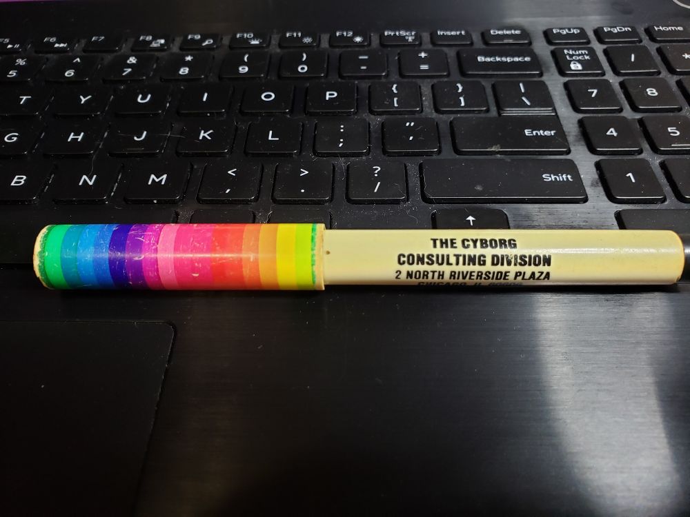 A beige pen sits on a black laptop keyboard. On the cap is a rainbow of colours, on the barrel reads:
THE CYBORG 
CONSULTING DIVISION
2 NORTH RIVERSIDE PLAZA