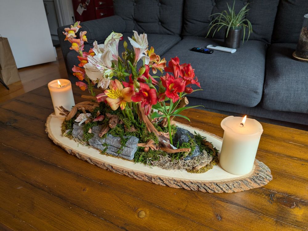 A photo of a table centerpiece on a coffee table. The centerpiece has a thin slice of a log as the base, with stones, driftwood, and preserved moss glued into a natural scene on top. There are flowers coming out of the scene, and two candles on either side of the centerpiece.