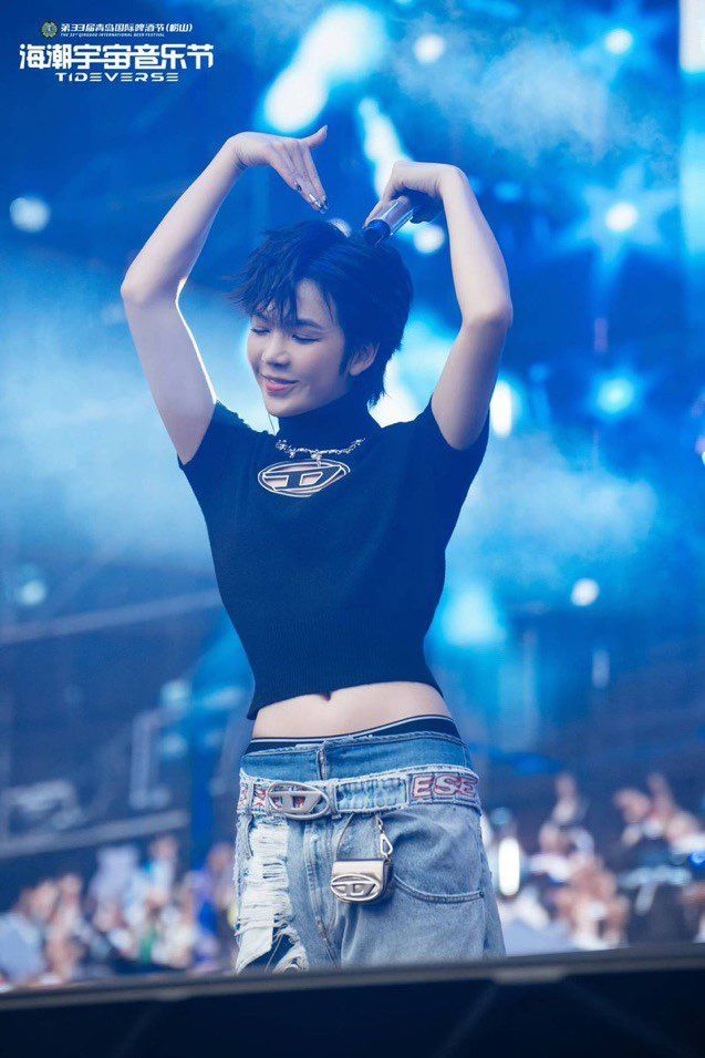 Thai actor Earth aka Cooheart on stage at a fan event. He is holding his arms above his head to make a heart shape. This has caused the shirt he is wearing to rise up, emphasizing his waist and showing just enough of his stomach you can see his belly button.
Amidst the bright lights and lens flare around him, he is serene.