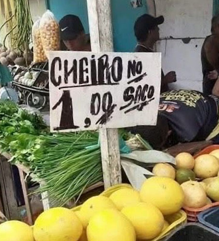 Imagem de uma barraca de feira com frutas e verduras. Sobre o cheiro verde, uma plaquinha: CHEIRO NO SACO R$ 1,00