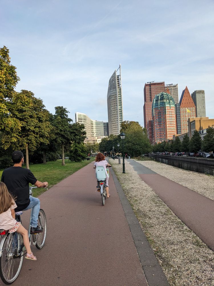Vater, Mutter, Kind auf drei Fahrrädern auf rot asphaltierter Radinfrastruktur. Im Hintergrund die Skyline von Den Haag