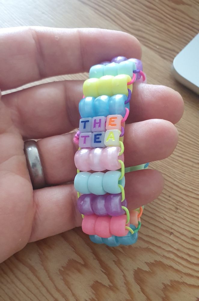 A hand is holding a multicolour bead bracelet. Within the bracelet, the words "THE TEA" are visible.