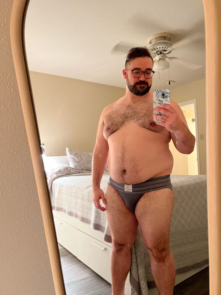 The poster stands at the foot of his bed in a new, dark gray jockstrap, featuring a wide, striped waistband. Otherwise, he stands nude in a full length mirror. 