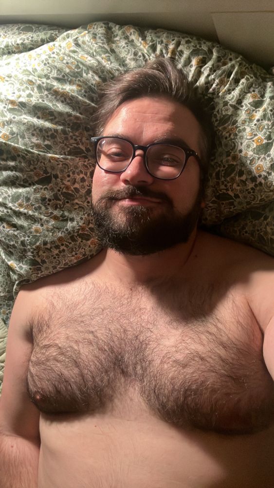 The subject looks into the camera with a sleepy look in his eyes, still wearing his glasses in bed, although he’s clearly ready to fall asleep. His chest and some torso are also exposed, revealing hairy pecs and nipples. 