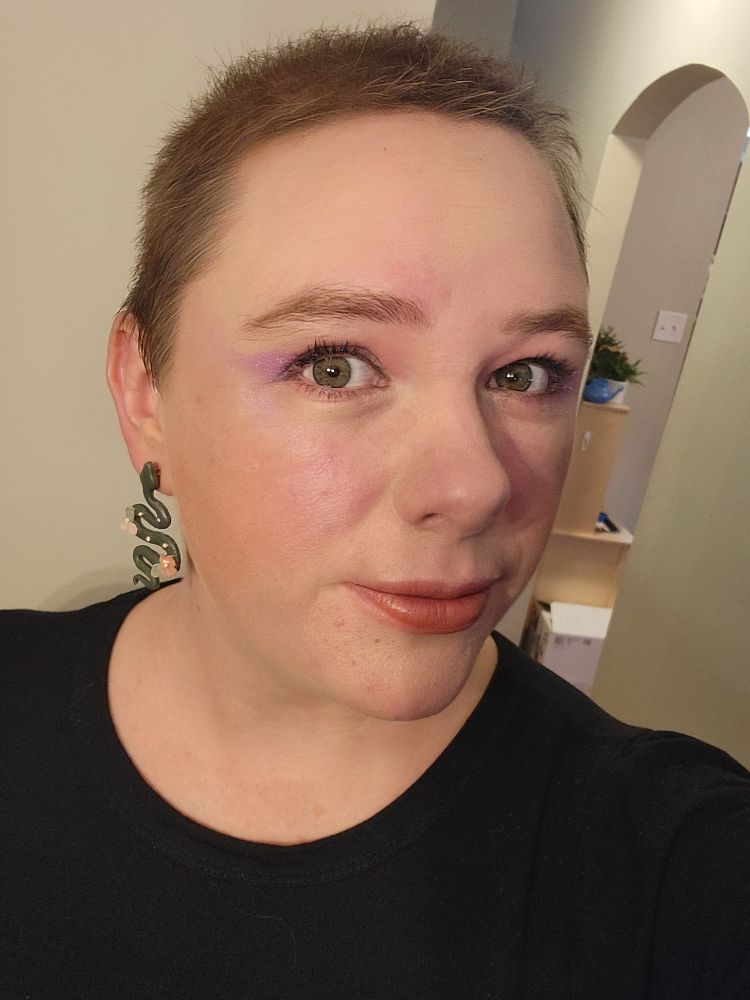 Color photo of white woman with short brown hair. She has a black shirt on with snake earrings, with purple eye makeup and a berry red lip.