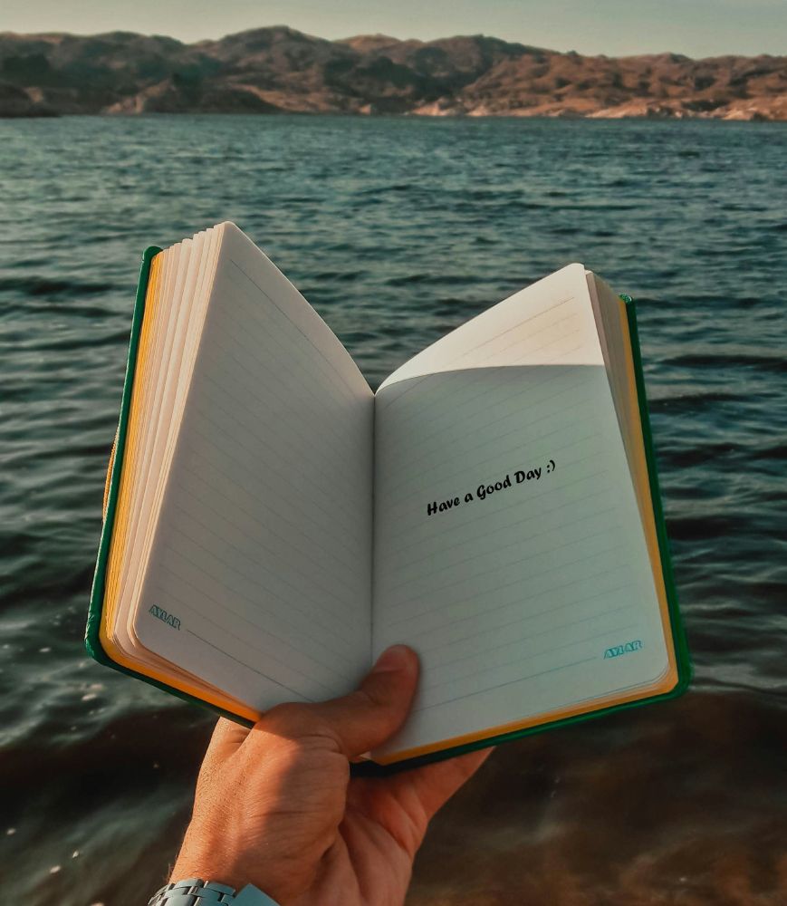 Somebody holds a book out near a lake. The book says Have a Good Day.