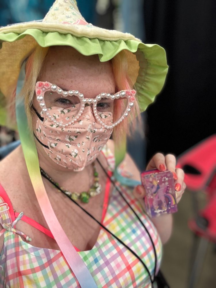A woman with short fluffy yellow & pink hair wearing a flowered pink kn95 mask, heart shaped pink glasses with pearls and pink roses all around the frames, an ivory felt witch hat with pastel flower sequins, rainbow pastel ribbons, and a mint green ruffled brim, a green glass beaded necklace, and a pastel rainbow gingham v-neck top. She is holding up a translucent rectangle soap with a neon pink & purple base and a Twilight Sparkle alicorn figurine embedded inside. 