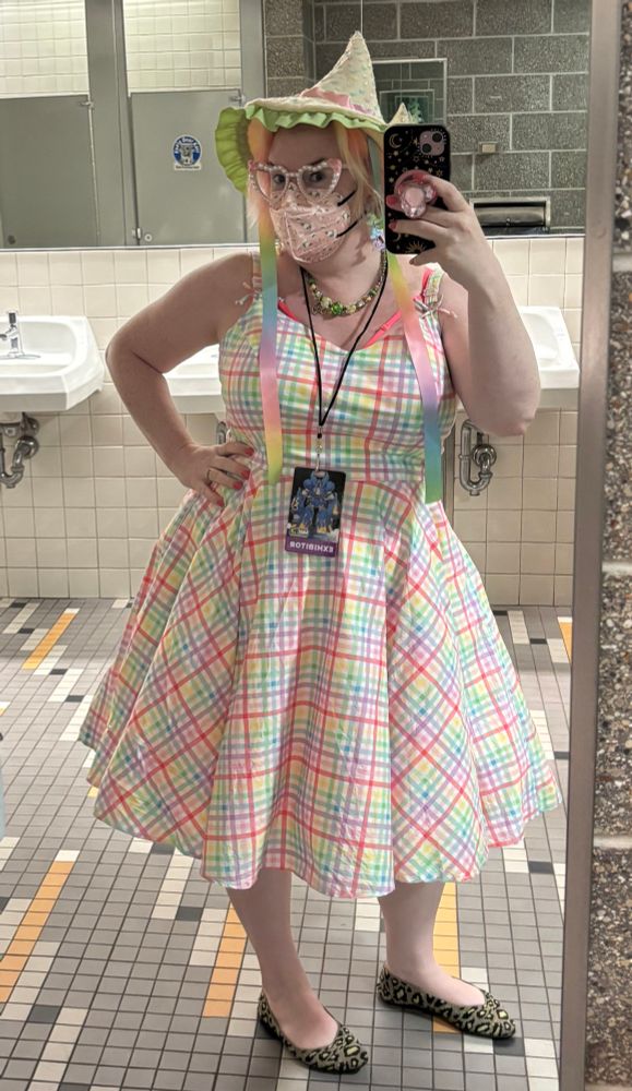 Bathroom mirror shot of a woman in a rainbow pastel gingham swing dress with straps & poofy skirt, a cream felt witch hat with pastel rainbow sequins and ribbons hanging down to the sides, heart shaped pearl encrusted glasses, a pink kn95 mask, a con badge on a black lanyard, and olive green leopard print flats. She has one hand on her hip and is taking the photo with her phone in the other hand. 