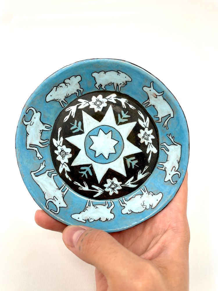  A blue, white, and black ceramic dish with a  barn quilt interior surrounded by flowers. The outer rim is lined with little, simply stylized sheep and cows.