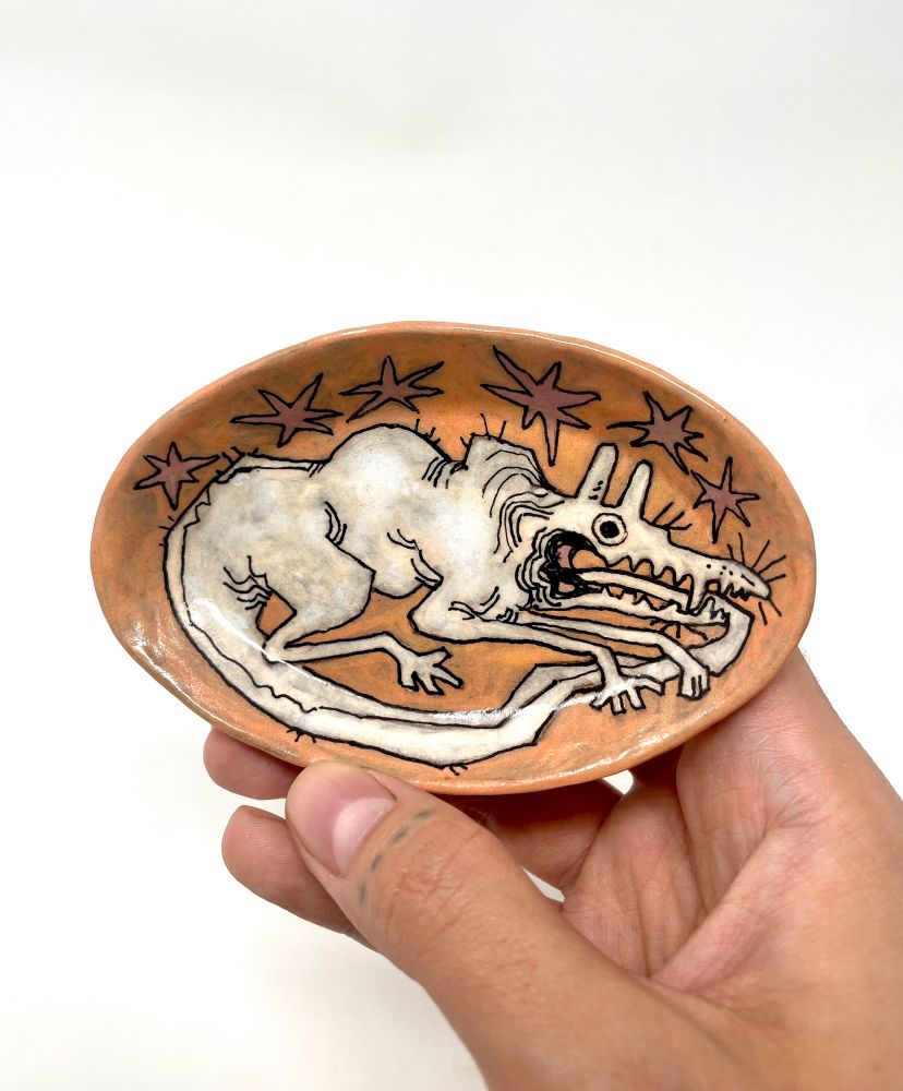 An oval shaped, peach coloured, ceramic dish. A white, wide eyed creature is lying, front paws folded over its tail, which descends into its open, toothy mouth, swallowing its tail.
