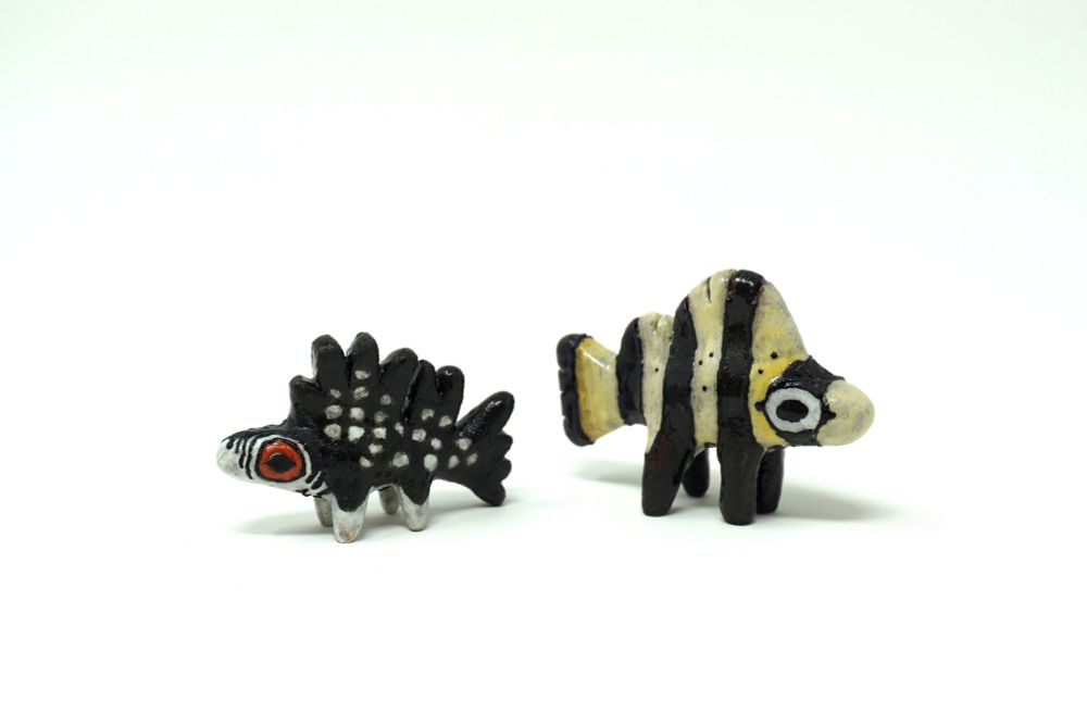 To the left, a ceramic and fictitious spiky finned and compact fish with legs. A black body contrasts a white underside. White spots dapple its body and its eyes a red-orange. To the right, a ceramic barred knifejaw with legs; a vertically striped black and white fish with hints of yellow against its cheeks and tail.