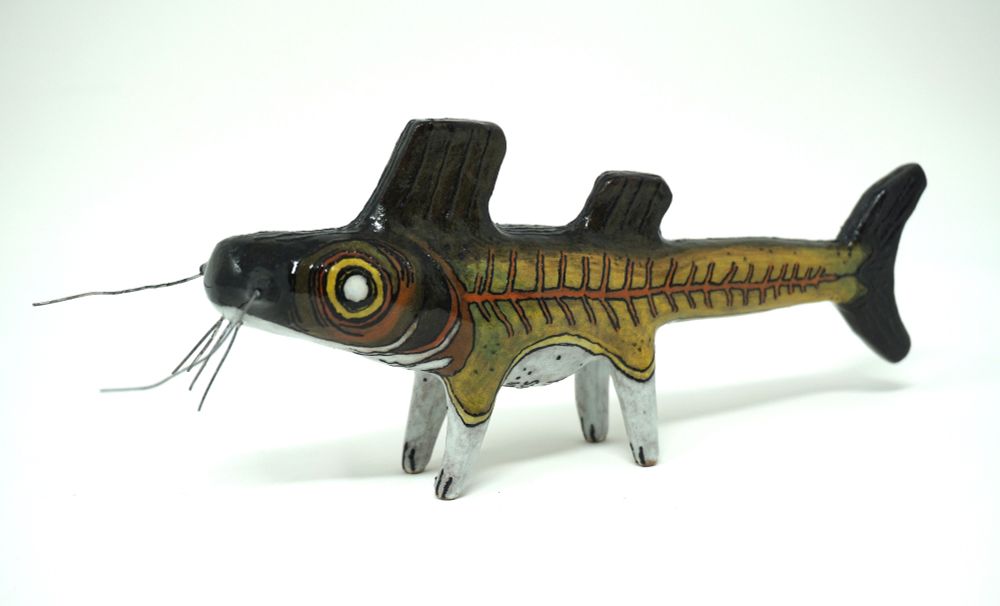 A ceramic catfish with legs. Its barbels are made out of wire. Its eyes are big and peering, the body is olive in colour with the very top half of its body and head black, and a pale white belly and legs. The skeleton like lateral line on its body is a rusty brown.