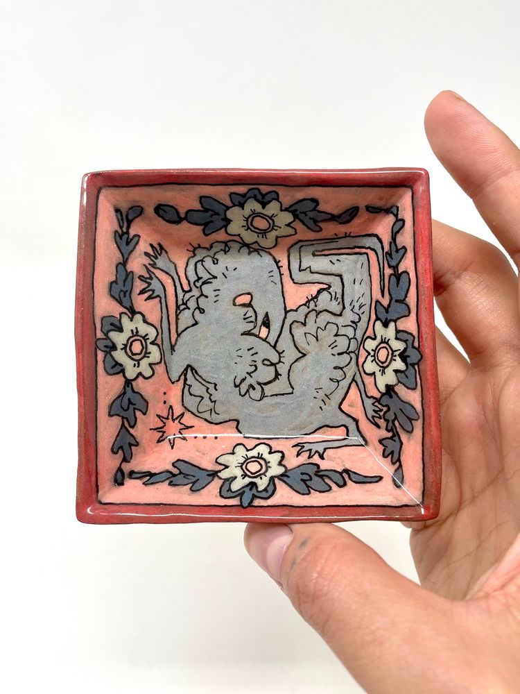 A soft pastel pink and blue square ceramic dish with a red rim. A fluffy creature is surrounded by flowers and its long neck loops over its back, akin to a bird, to bury its head in its fluffy, wooly coat. It shyly, perhaps sleepily, squints back at the viewer.