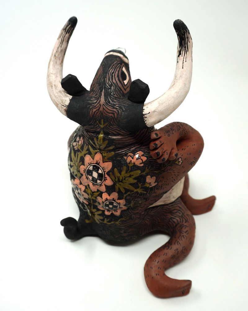 Multiple photographed angles of a ceramic sculpture of a fat, transmasc minotaur with a septum ring sitting on the ground, head thrown back, content and proud, locking eyes with the viewer. One hand sits on the ground, the other raised gently towards the neck. His body is a warm reddish-brown, with a white throat, chin, and belly, with a chest featuring scars from top surgery. Dense body hair gradually gives way to thick black fur covering his head, neck, back, and tail. Long horns tipped in black frame his face.
On his back sits a cluster of 2D illustrated flowers with pink petals, leafy green stems, and an interior consisting of a black and white checkerboard pattern. 