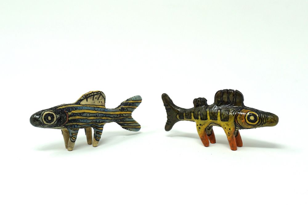 To the left, a ceramic zebra danio with legs; a slender blue and yellow striped fish. To the right, a ceramic yellow perch with legs; a green fish with darker barring on its body, the green fading to a warm yellow by its belly, and orange by its legs.