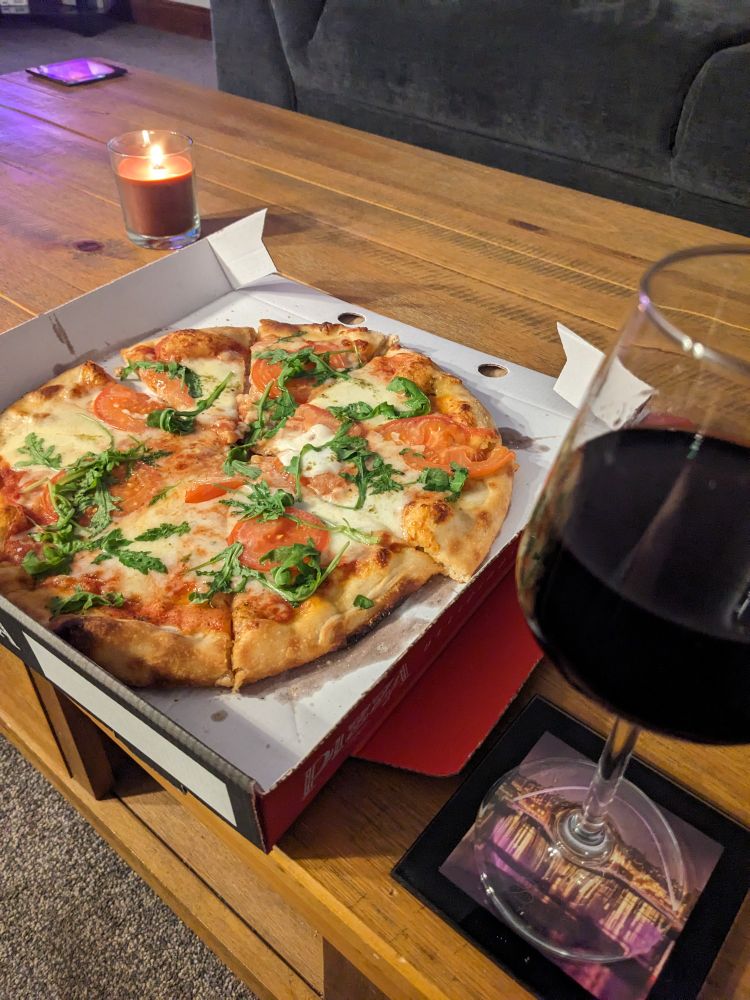 A pizza caprese sits in an open takeaway box on a wooden coffee table, in front of a grey sofa. The pizza has tomato, mozzarella, and rocket on top. A large glass of red wine is also in view.