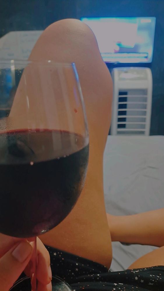eu estou relaxada na cama, segurando uma taça cheia até a metade de vinho tinto bordô, com uma televisão e um ventilador no fundo.