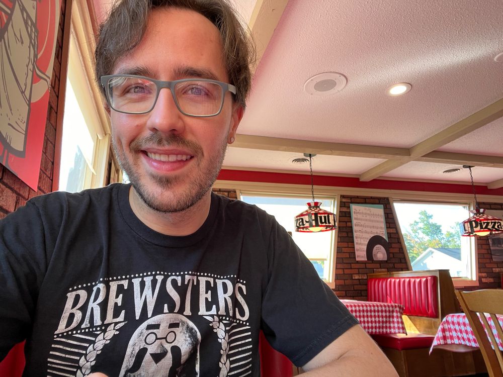A selfie taken in a classic Pizza Hut. The booths are a dated (yet new in quality) red color with red and white gingham table sloths. The lights above the table have the vintage Pizza Hut stained glass lampshades. The walls are a nice brick with various retro Book It program ad posters and photos of original Pizza Hut locations. Unpictured but they had a functional and pretty alright salad bar!

I have medium length brown hair that is starting to catch a wave. I’m wearing gray classes and a black Brewster’s shirt (Animal Crossing). This was post salad and cheesy garlic breadsticks but before stuffed crust pizza. 