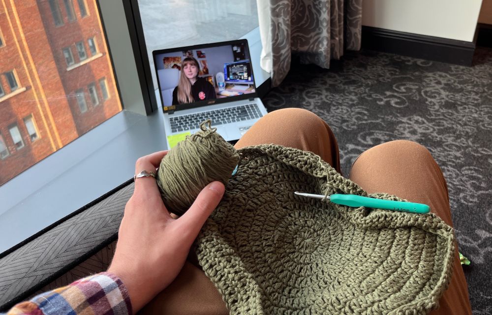 POV shot of me sitting on a hotel chair overlooking a river. On my lap is a green coboo yarn beanie about 2/3 of the way completed. On the window sill is my laptop playing a YouTube video about the indie web. I’m wearing a yarn tension ring as I’m still recovering from my latest arm surgery but am finally able to get back to work on my crochet a little at a time. You can see my orange-brown hiking pants and orange/blue/burgundy flannel sleeve. It’s almost fall and my outfit is celebrating early. 