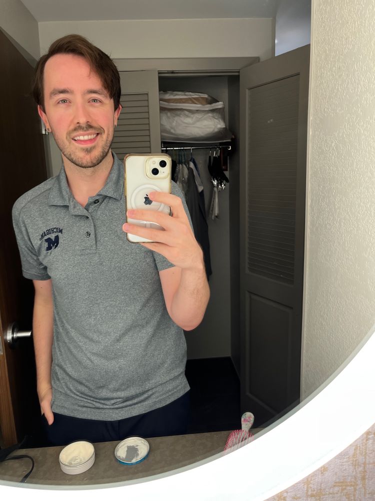 Me with a lightly trimmed beard and medium length brown hair stands smiling in a hotel mirror in a grey polo with Michigan and an M embroidered in dark blue on the top right. 

The closet door behind me is open revealing a pillow and some of our dress shirts suit jackets hung up. An open styling clay container is open on the counter next to a hair brush. The mirror is entirely framed in a ring light. If I had aesthetic sense I would’ve cleaned all of that up before taking the photo but I was enraptured in the lighting. Gay moth to the flame and all of that. 