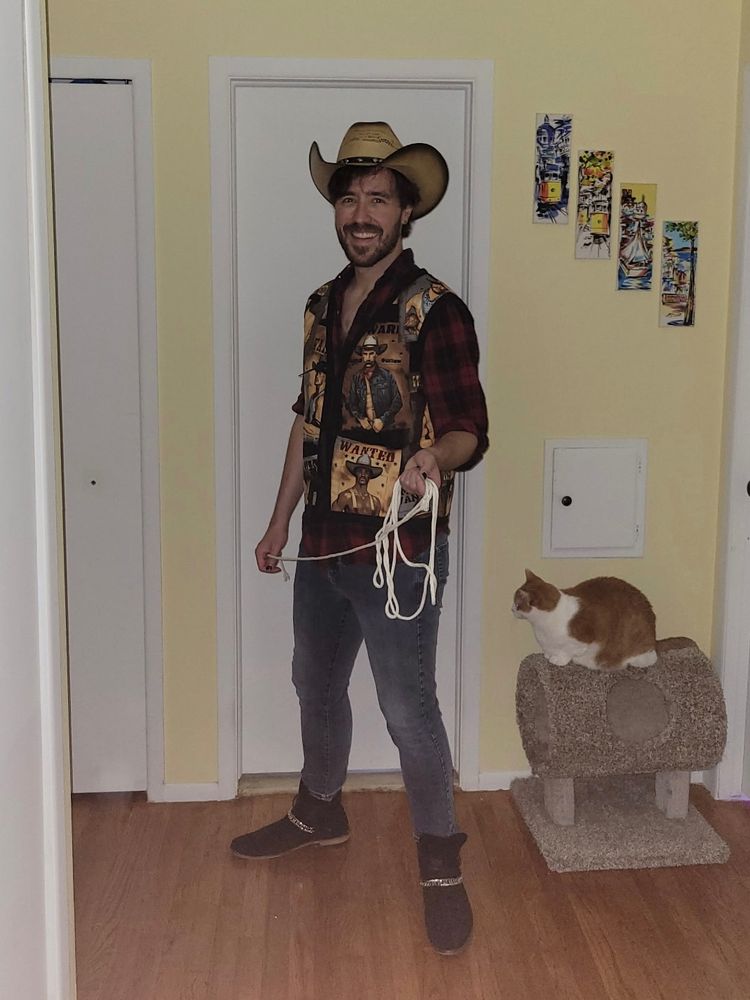 I stand uncovered in a slutily unbuttoned red flannel with me made gay cowboy vest, cowboy hat, and black cowboy boots with silver chains. I’m holding some crafting rope like a lasso and the photo was shot with flash in the dark. My cat is on a cat tree next to me paying me no mind. 