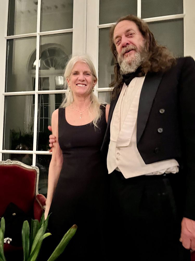 Woman and man dressed in formalwear big smiles in front of large paned windows. 