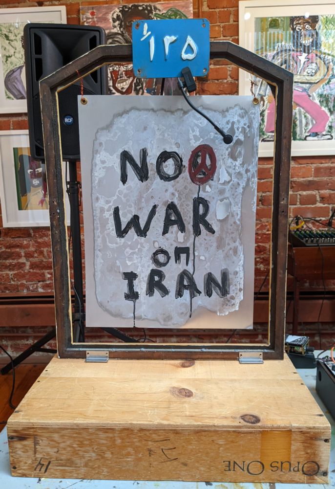 Text on a metal plate suspended on a wooden frame: NO WAR ON IRAN
