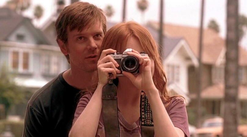 Capture de la série Six feet Under où le personnage de Nate Fisher (Peter Krause) chuchote à l'oreille de sa sœur Claire Fisher (Lauren Ambrose) qui prend une photo.