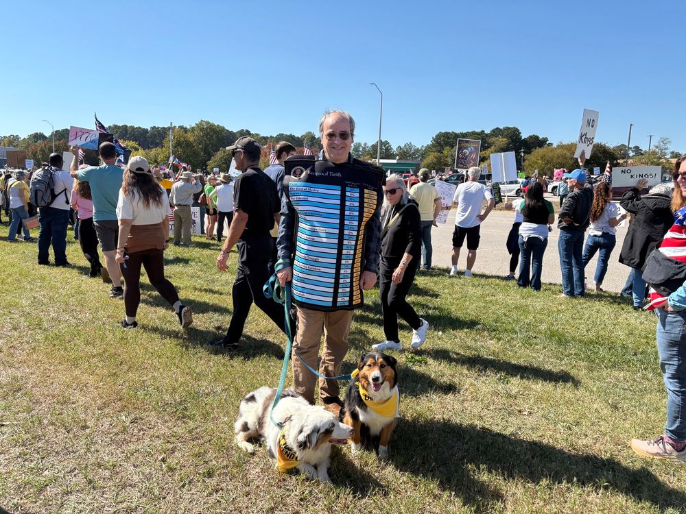 A man wearing a costume with a list of tariff rates stands beside two dogs.