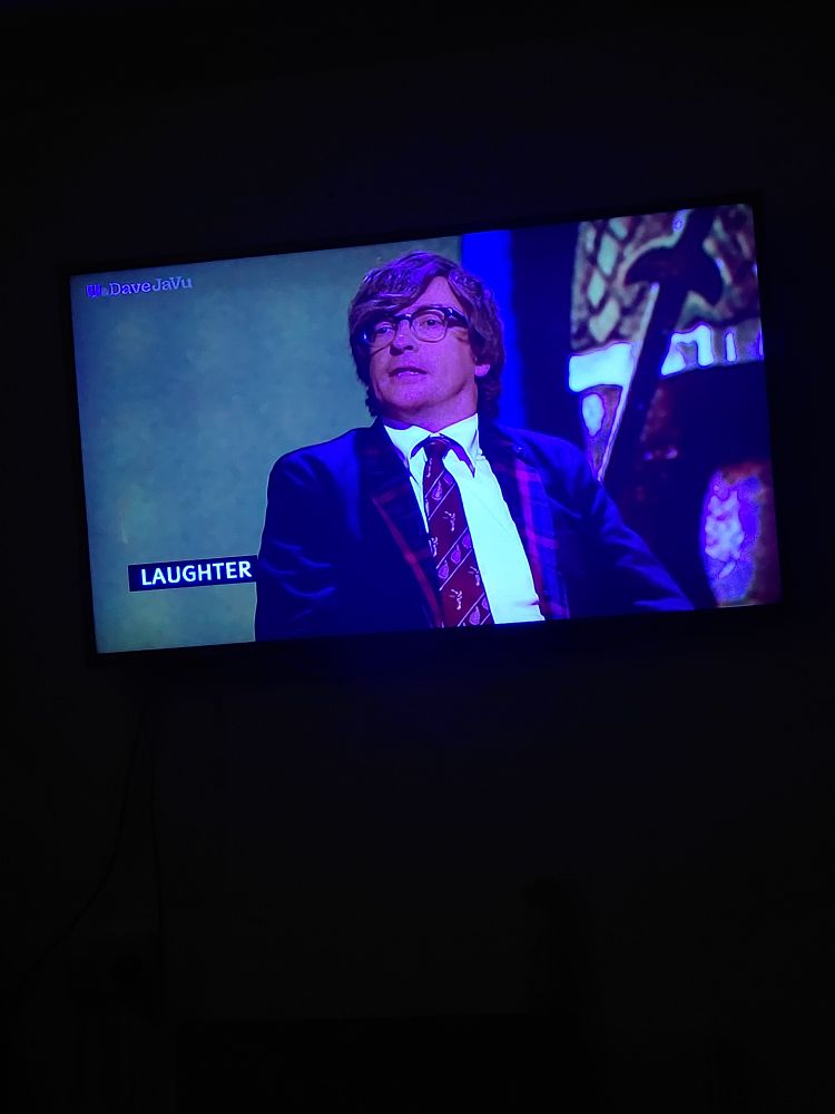 Photo of Rhys Darby on TV taking part in QI. He is wearing a suit, red tie, white shirt and glasses. Closed caption "Laughter"