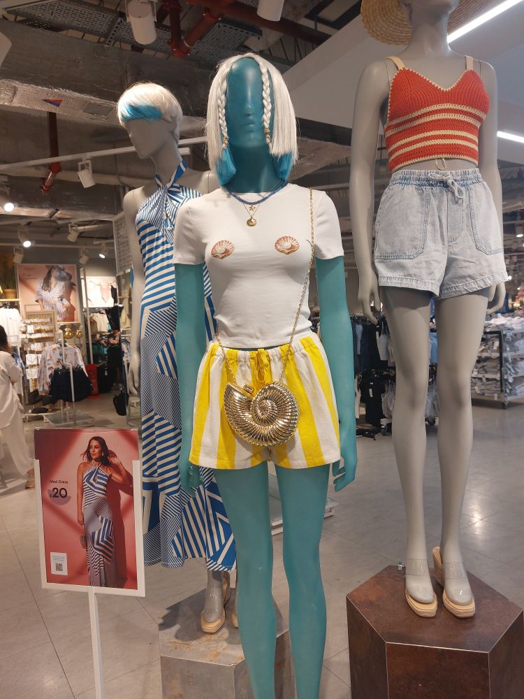 Photo of Primark mannekins wearing summer clothing.

Left - cropped blonde wig with patch of blue dye, blue stripey maxi dress with a photo of a model wearing the dress in front.

Middle - blonde bob wig with two plaits to frame the face, white t-shirt with two shells on across the the breasts, stripey white and yellow shorts and golden shell cross body bag.

Right - woven wide brim summer hat, knitted yellow and orange vest top and demon shorts). 