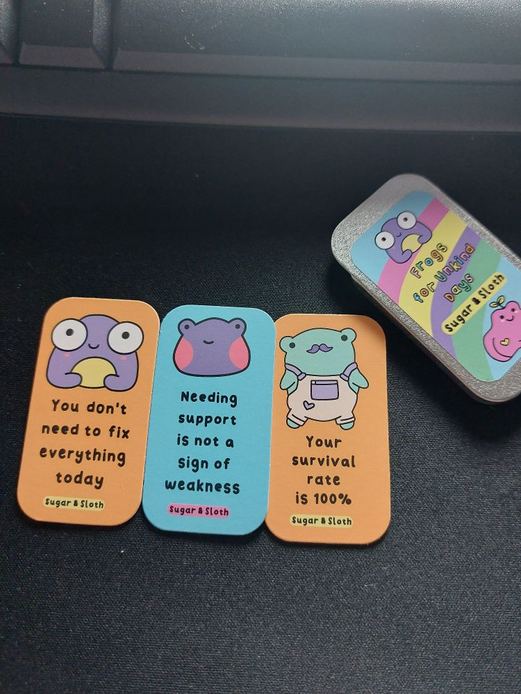 Picture of tiny affirmation cards with cute frogs by Sugar & Sloth. 
Left to right: orange card with purple frog says: "you don't have to fix everything today".
Blue card with purple frog says: "needing support is not a sign of weakness".
Orange card with green frog wearing beige dungarees and has a purple moustache says: "your survival rate is 100%".
The right hand tin lid has a sticker with a rainbow background, a purple frog and a pink frog. It says: "Frogs for Unkind Days" in rainbow lettering. 