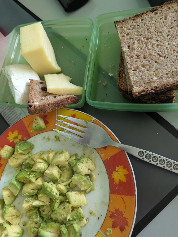 Teller mit grünen Avocadostücken und Gabel. Darüber grüne Tupperdose mit braunem Brot und Käsestücken.